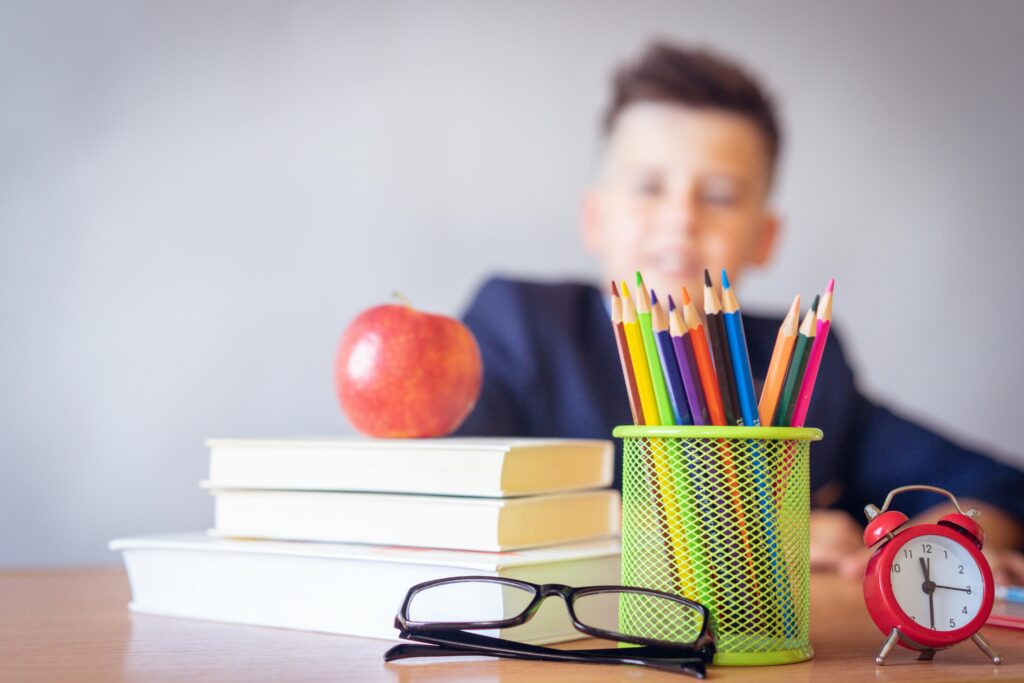 pexels-photo-2781814-2781814-1 Schoolboy smiling behind a desk with books, pencils, and an alarm clock symbolizing study and creativity.