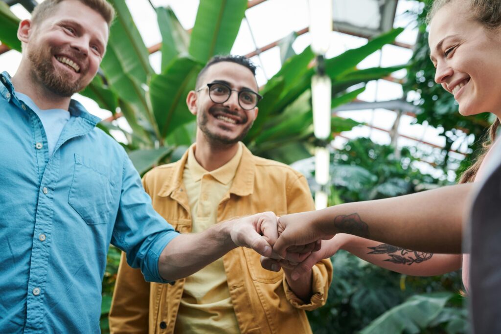 pexels-photo-3228690-3228690 Three friends sharing a joyful fist bump in a lush indoor greenhouse environment.